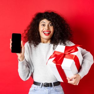 valentines lovers day excited smiling woman with curly dark hair showing smartphone empty screen holding surprise gift holiday showing online promo red background x
